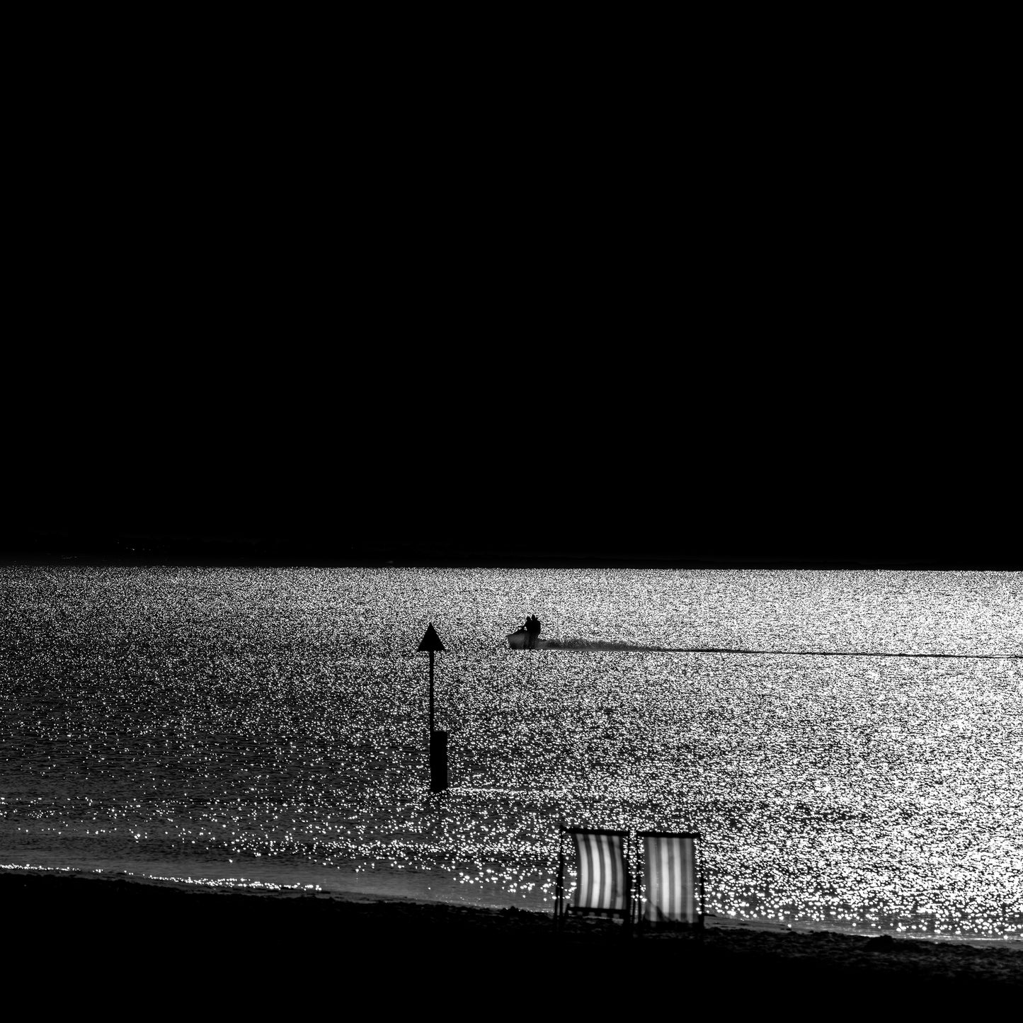 Isu to Umi — two empty deckchairs on dark shore with jet ski on glittering silver sea, black and white fine art photography