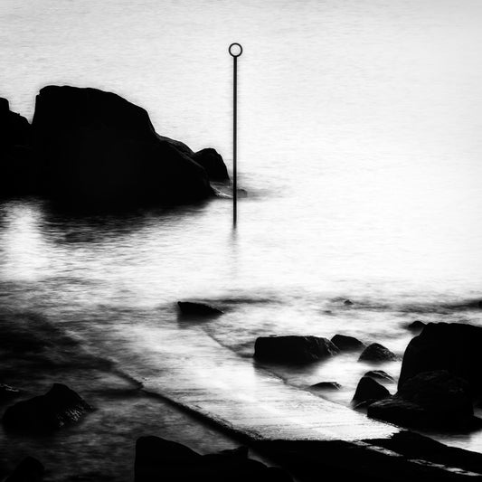 The Marker — slender pole with circle top standing in luminous misty long-exposure water with dark rock silhouette and scattered stones, black and white fine art photography by Roy Fraser