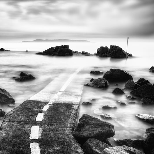 This Way — dashed white line on a stone slipway leading into misty long-exposure sea with rocks and marker pole, black and white fine art photography by Roy Fraser