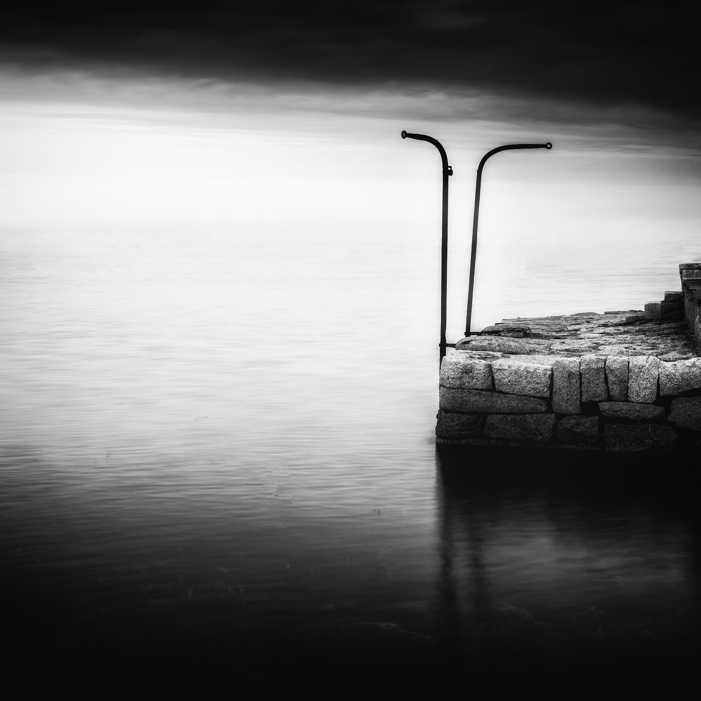The Gatekeepers — two iron posts with curved tops on a granite jetty reflected in still long-exposure water under a luminous horizon, black and white fine art photography by Roy Fraser