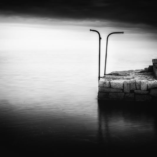 The Gatekeepers — two iron posts with curved tops on a granite jetty reflected in still long-exposure water under a luminous horizon, black and white fine art photography by Roy Fraser
