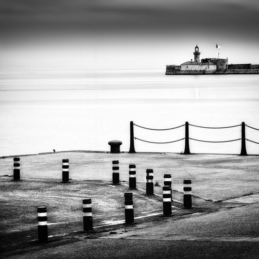The Watch — harbour quayside with striped bollards and chain-linked posts, lighthouse and fortified building on the horizon across a silky long-exposure sea, black and white fine art photography by Roy Fraser