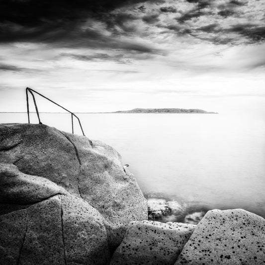 The Way Down — triangular iron railing atop a granite boulder with silky long-exposure sea and distant island under a brooding sky, black and white fine art photography by Roy Fraser
