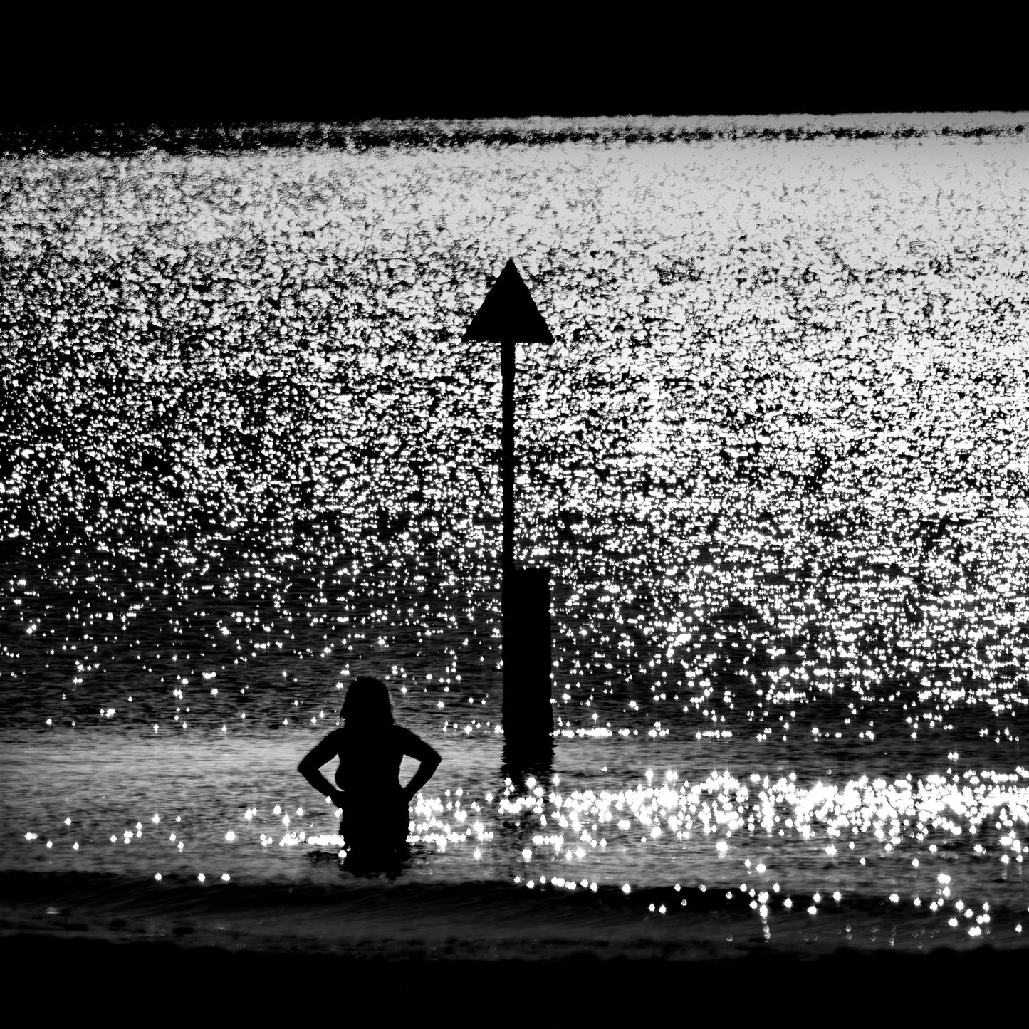 Chinmoku no Kishi — woman waist-deep in sparkling sea beside triangular beach marker, black and white fine art photography