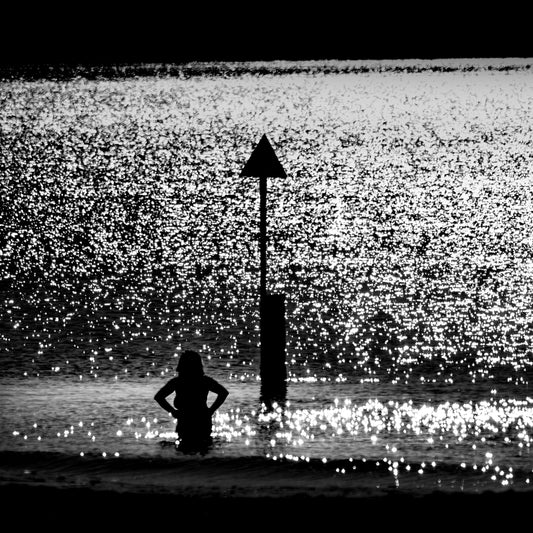 Chinmoku no Kishi — woman waist-deep in sparkling sea beside triangular beach marker, black and white fine art photography