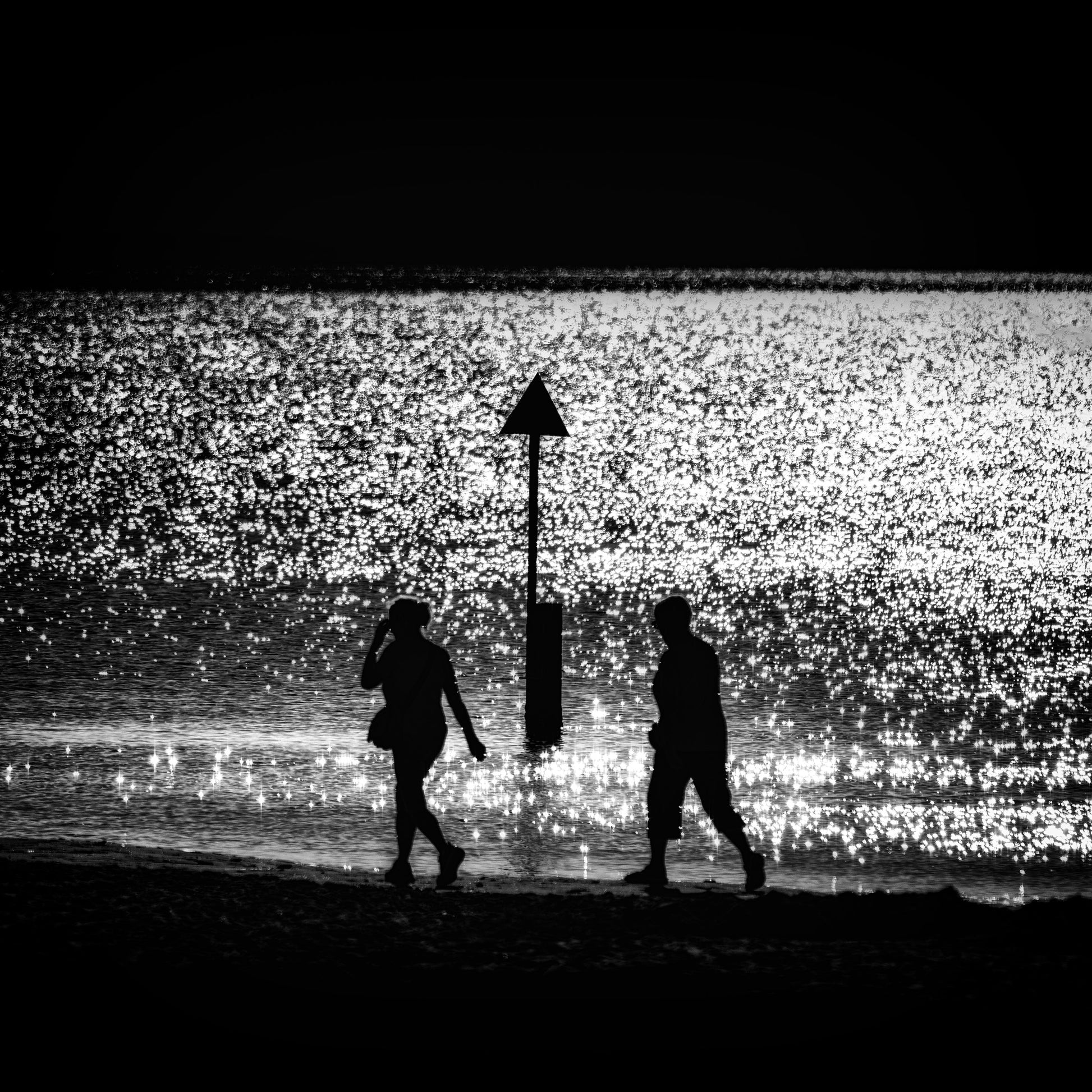 Futari no Kishi — couple silhouetted walking either side of triangular beach marker on glittering shoreline, black and white fine art photography
