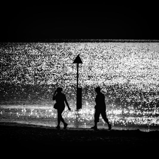 Futari no Kishi — couple silhouetted walking either side of triangular beach marker on glittering shoreline, black and white fine art photography