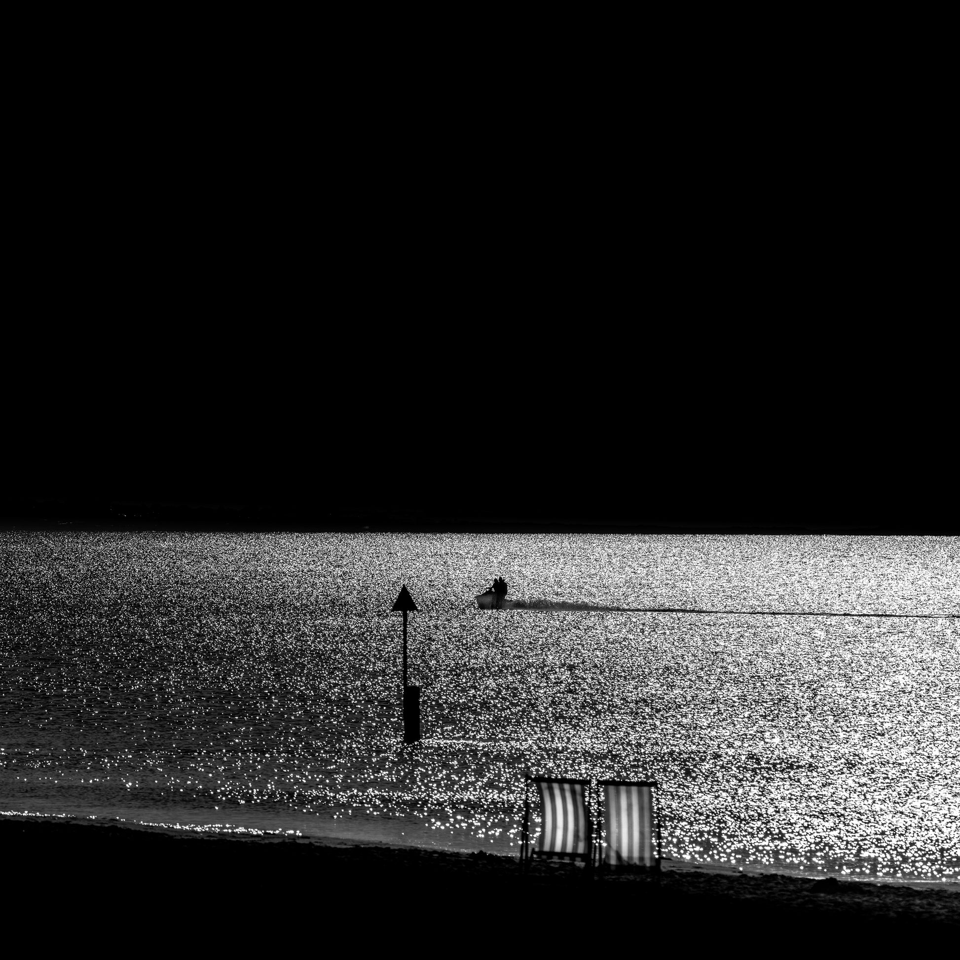 Isu to Umi — two empty deckchairs on dark shore with jet ski on glittering silver sea, black and white fine art photography