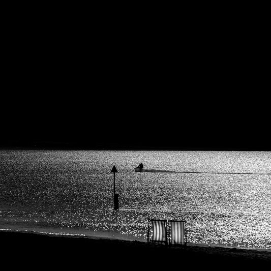 Isu to Umi — two empty deckchairs on dark shore with jet ski on glittering silver sea, black and white fine art photography