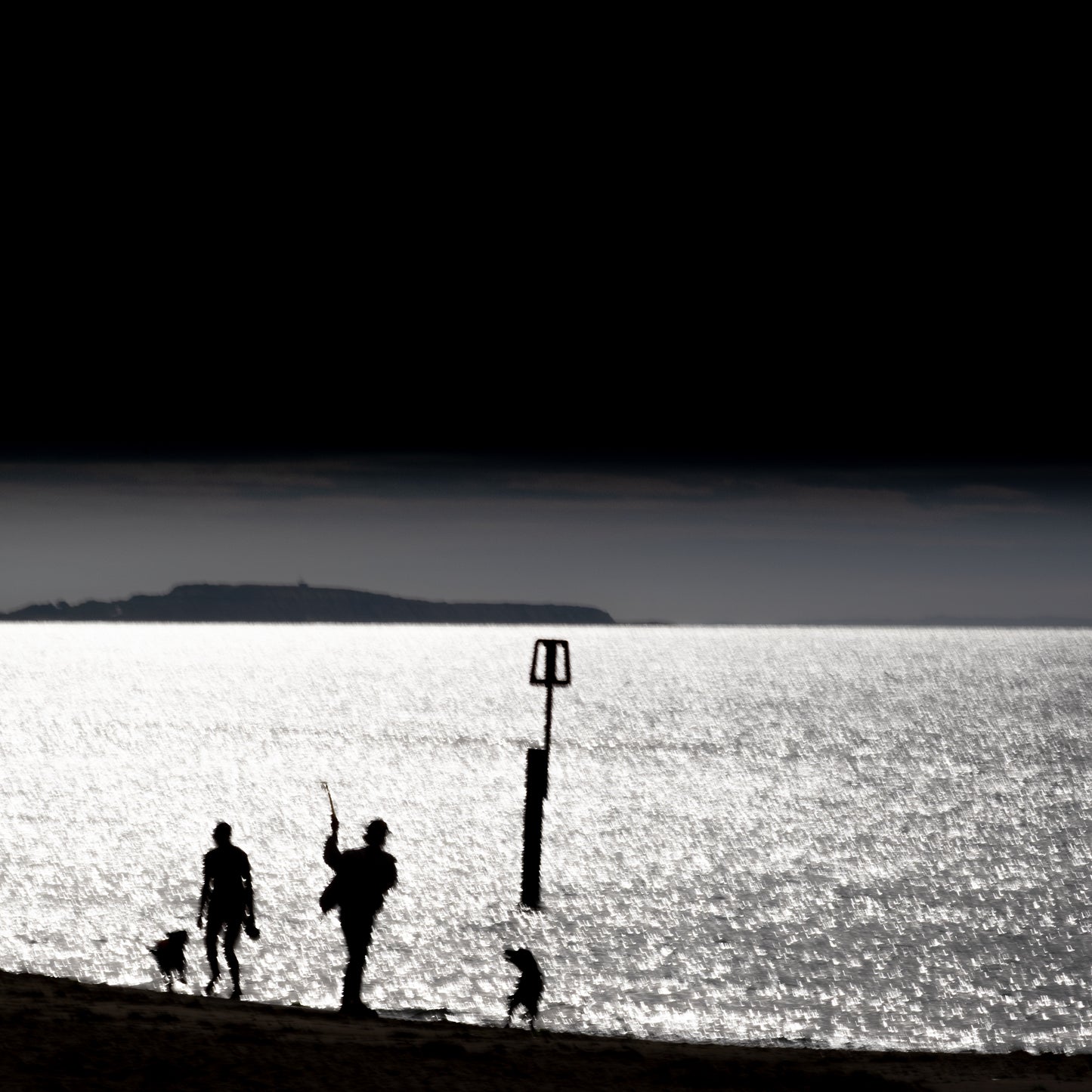 Kazoku no Nagare — family silhouette with dog on glittering shoreline, rectangular beach marker, island on horizon, black and white fine art photography