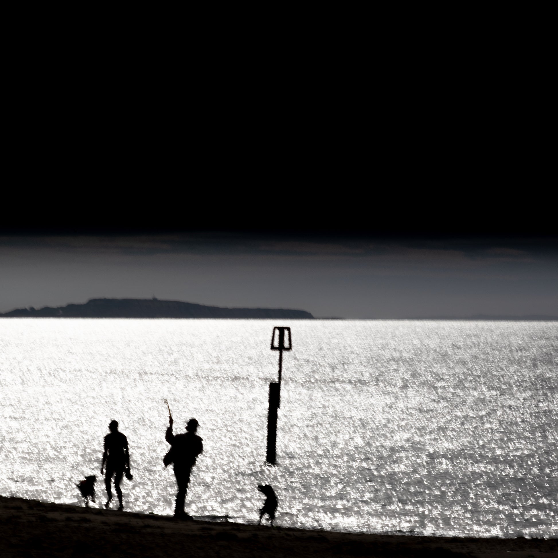 Kazoku no Nagare — family silhouette with dog on glittering shoreline, rectangular beach marker, island on horizon, black and white fine art photography